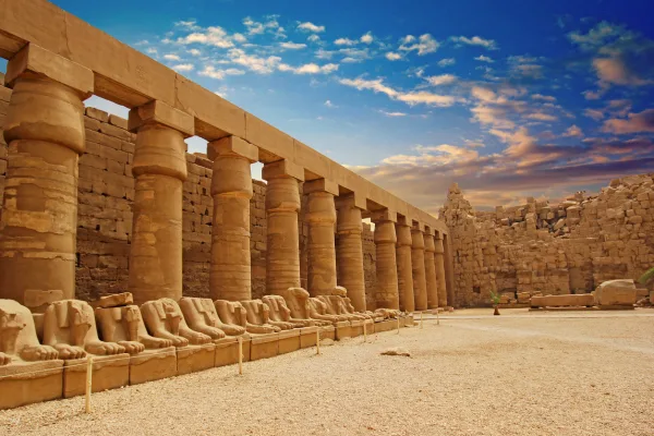 Ancient ram-headed sphinx statues lining the entrance to Karnak Temple in Luxor, Egypt, with towering stone columns against a blue sky