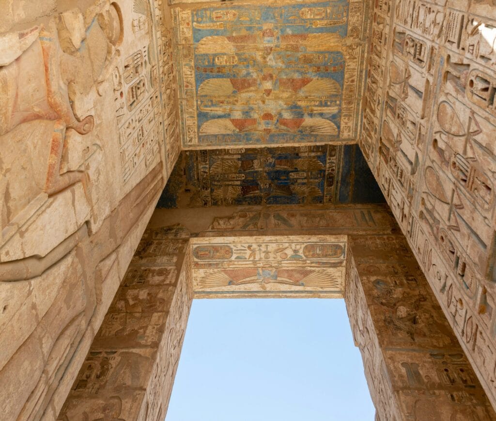 Looking up at the colorful painted ceiling and carved hieroglyphic walls of Medinet Habu temple in Luxor Egypt showing winged sun disks and ancient Egyptian reliefs