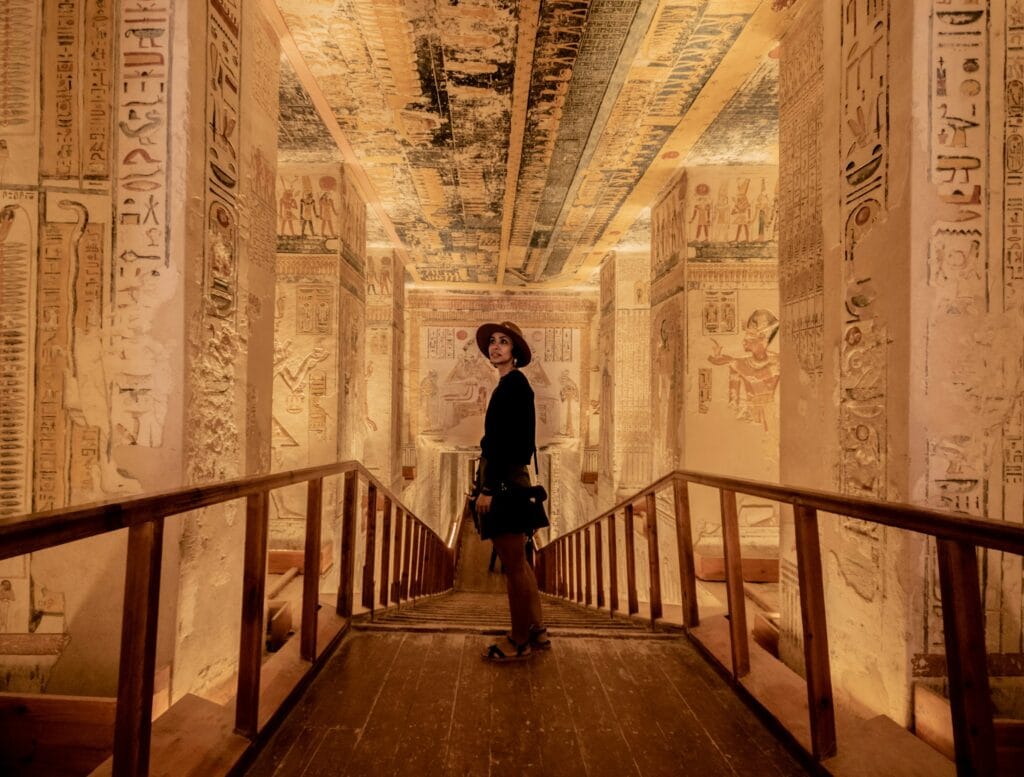 Visitor exploring a decorated corridor inside a royal tomb in the Valley of the Kings, Luxor, Egypt, with colorful ancient Egyptian hieroglyphics and painted figures covering the walls and ceiling