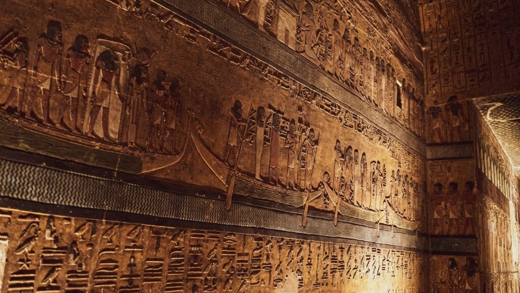 Ancient Egyptian tomb wall inside the Valley of the Kings showing carved and painted scenes of the solar barque carrying gods through the underworld with rows of hieroglyphics and figures