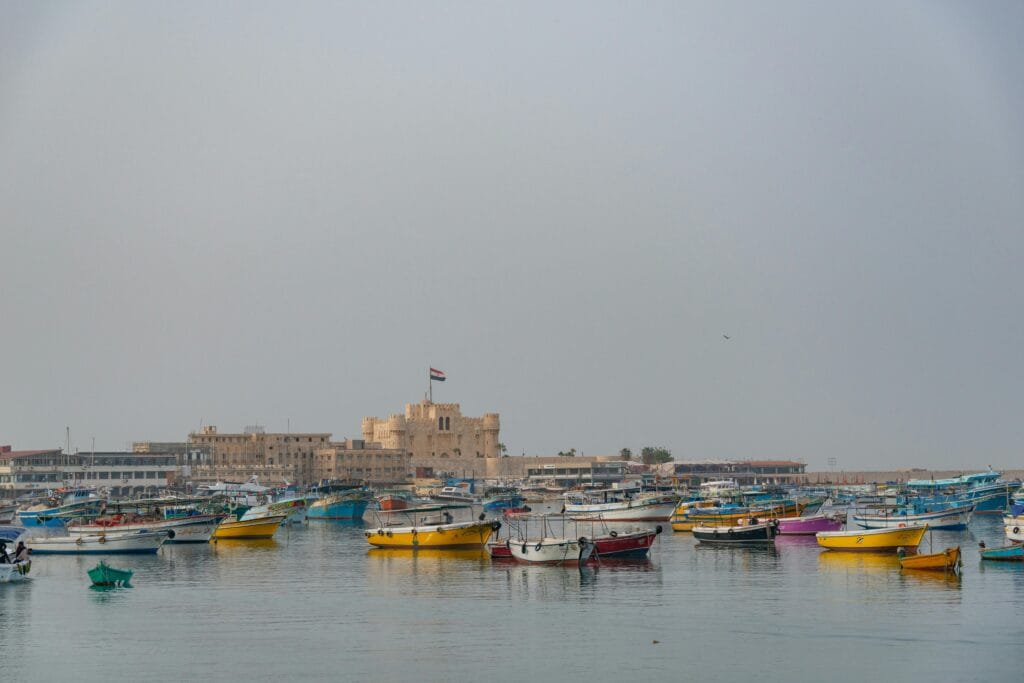 Colorful fishing boats anchored in Alexandria harbor with the historic Citadel of Qaitbay overlooking the Mediterranean Sea in Egypt.